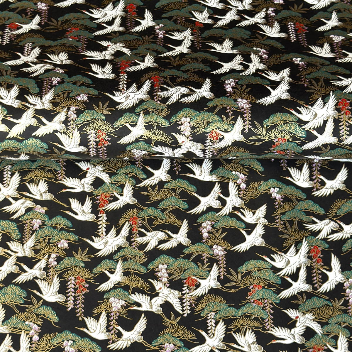 A close-up of a sheet of Japanese chiyogami paper featuring white cranes flying over green pine trees and branches on a black background.