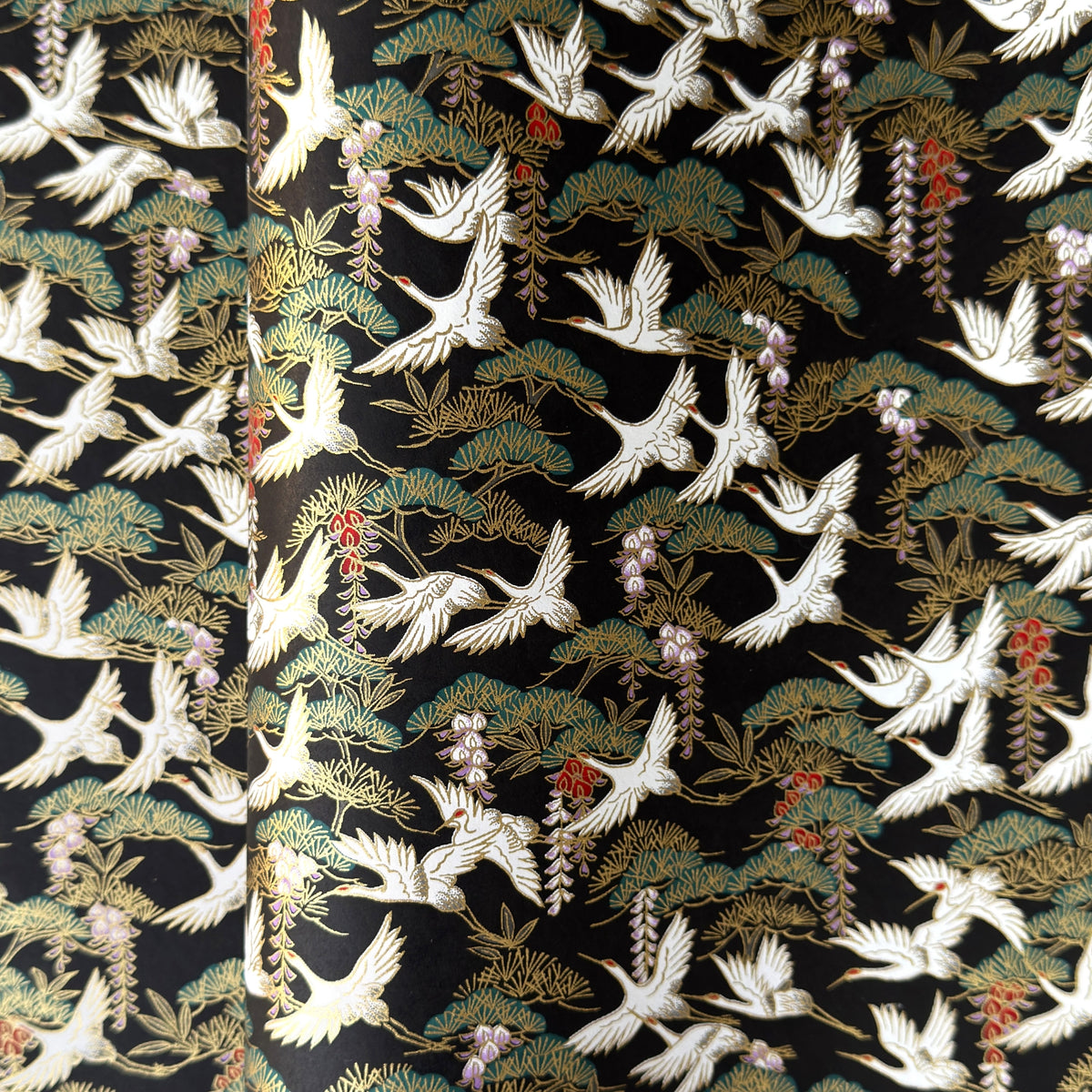 A close-up of a sheet of Japanese chiyogami paper featuring white cranes flying over green pine trees and branches on a black background.