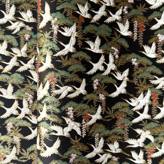 A close-up of a sheet of Japanese chiyogami paper featuring white cranes flying over green pine trees and branches on a black background.