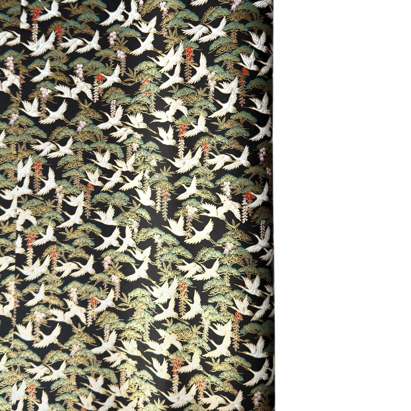 A close-up of a sheet of Japanese chiyogami paper featuring white cranes flying over green pine trees and branches on a black background.