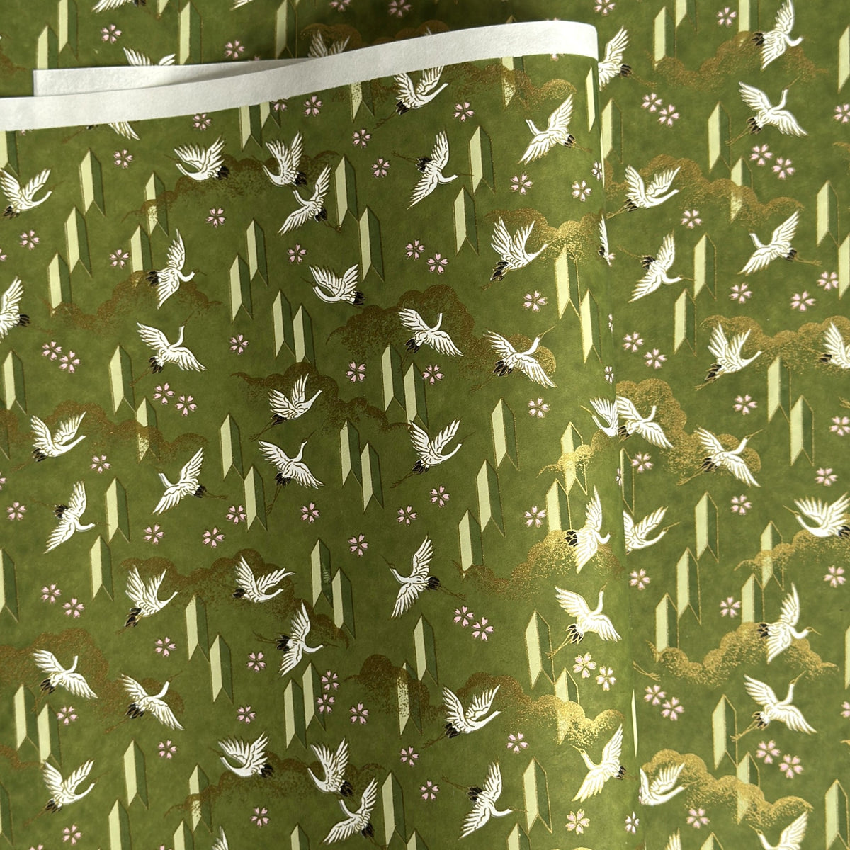 a sheet of Japanese chiyogami paper with an olive green ground and a pattern of white flying cranes and arrows.  There are also little pink cherry blossom flowers and gold clouds.