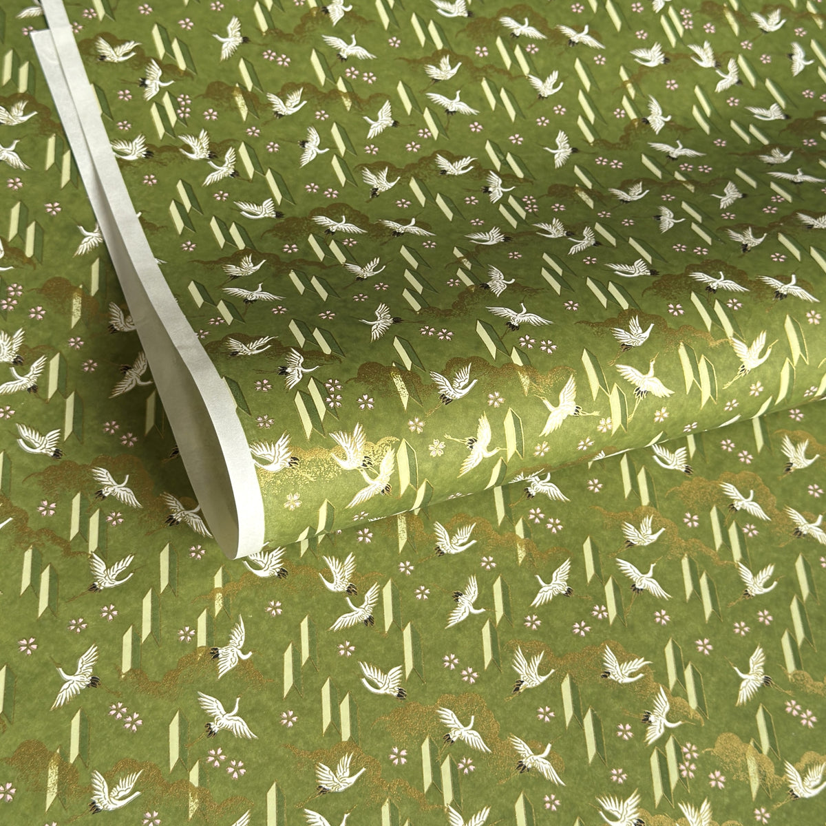a sheet of Japanese chiyogami paper with an olive green ground and a pattern of white flying cranes and arrows.  There are also little pink cherry blossom flowers and gold clouds.