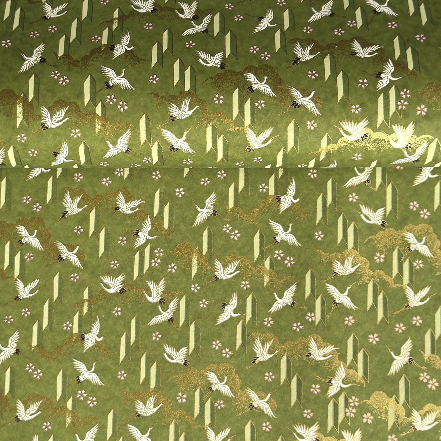 a sheet of Japanese chiyogami paper with an olive green ground and a pattern of white flying cranes and arrows.  There are also little pink cherry blossom flowers and gold clouds.