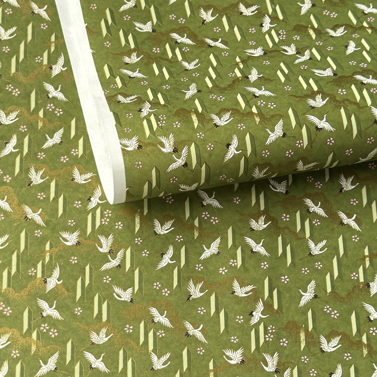 a sheet of Japanese chiyogami paper with an olive green ground and a pattern of white flying cranes and arrows.  There are also little pink cherry blossom flowers and gold clouds.