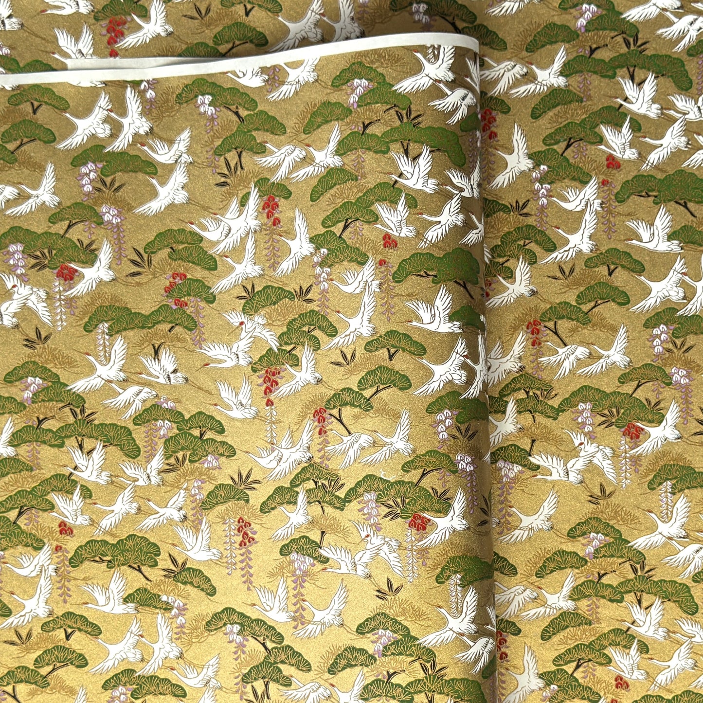 A close-up of a sheet of Japanese chiyogami paper with a repeating Japanese-style pattern featuring white cranes, green pine trees, and small red floral accents on a gold background.