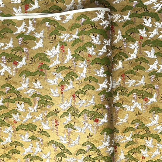 A close-up of a sheet of Japanese chiyogami paper with a repeating Japanese-style pattern featuring white cranes, green pine trees, and small red floral accents on a gold background.