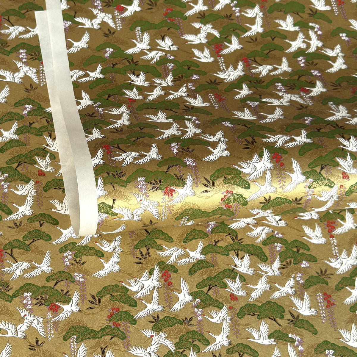 A close-up of a sheet of Japanese chiyogami paper with a repeating Japanese-style pattern featuring white cranes, green pine trees, and small red floral accents on a gold background.