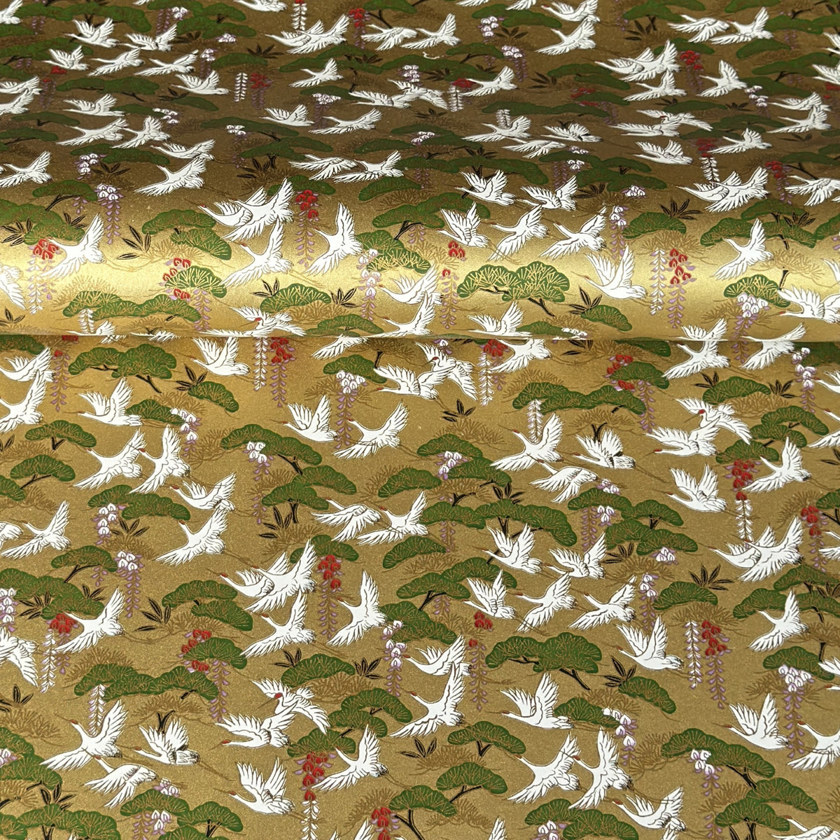 A close-up of a sheet of Japanese chiyogami paper with a repeating Japanese-style pattern featuring white cranes, green pine trees, and small red floral accents on a gold background.