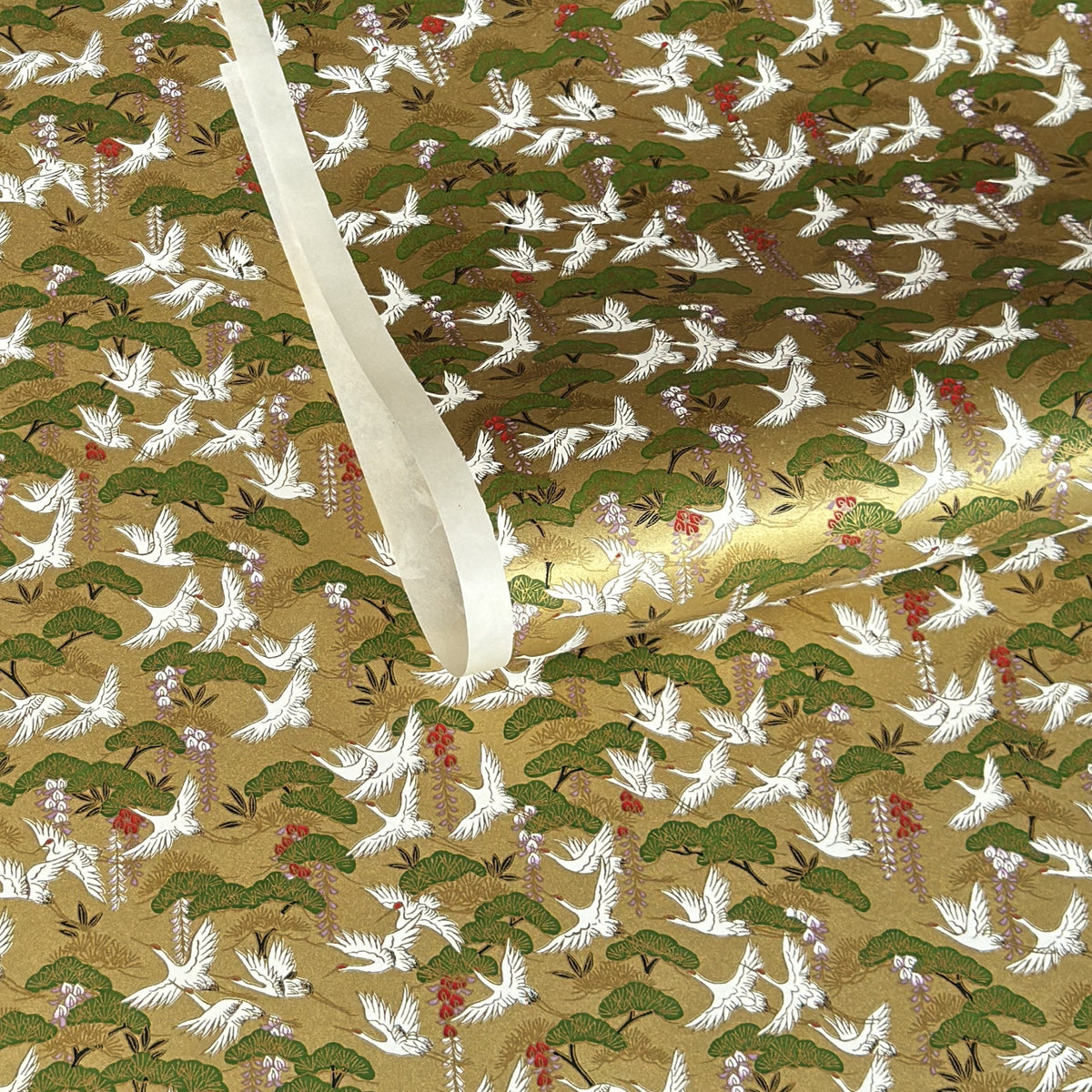 A close-up of a sheet of Japanese chiyogami paper with a repeating Japanese-style pattern featuring white cranes, green pine trees, and small red floral accents on a gold background.
