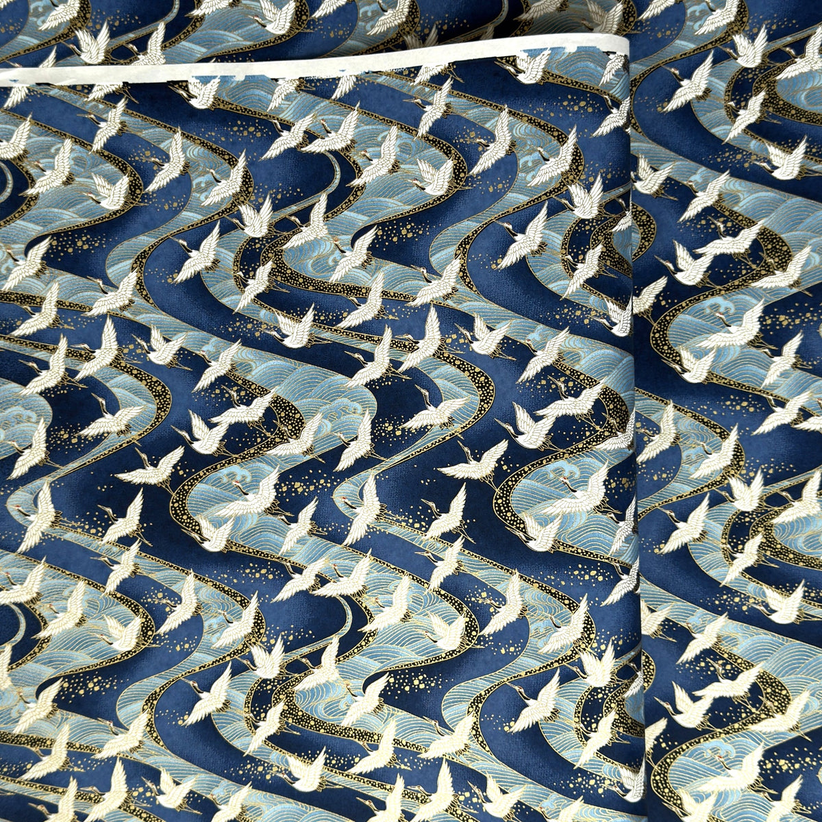 a sheet of Japanese Chiyogami paper with a hand screen printed pattern of white cranes in flight. The backdrop is swirling lines of dark and light blue with gold accents.