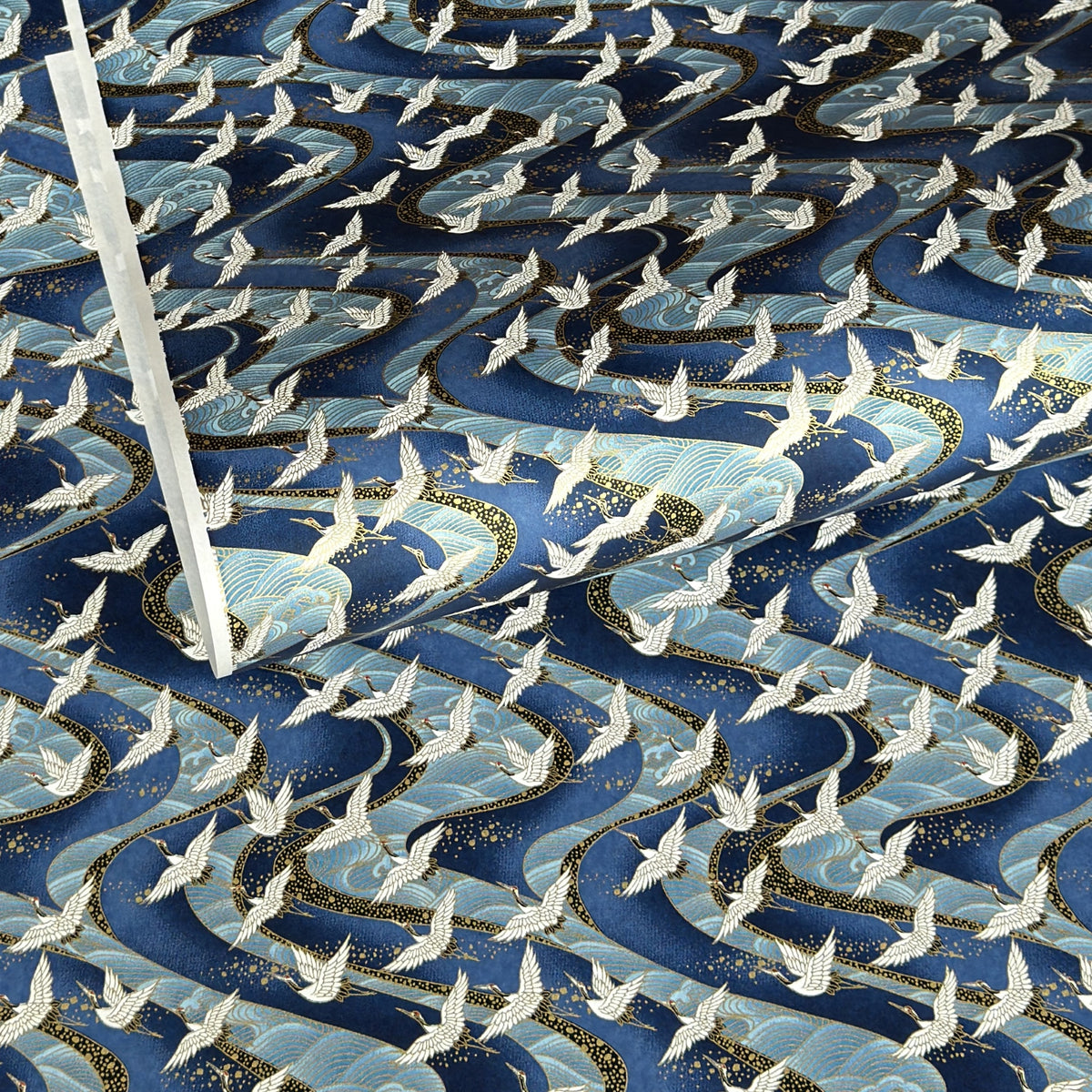 a sheet of Japanese Chiyogami paper with a hand screen printed pattern of white cranes in flight. The backdrop is swirling lines of dark and light blue with gold accents.