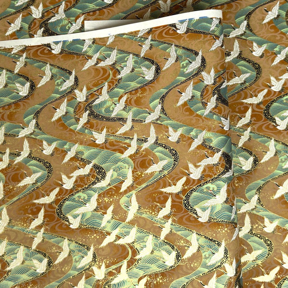 a sheet of Japanese chiyogami yuzen paper screen printed with a pattern of white flying cranes on a background of aqua and tan swirling lines