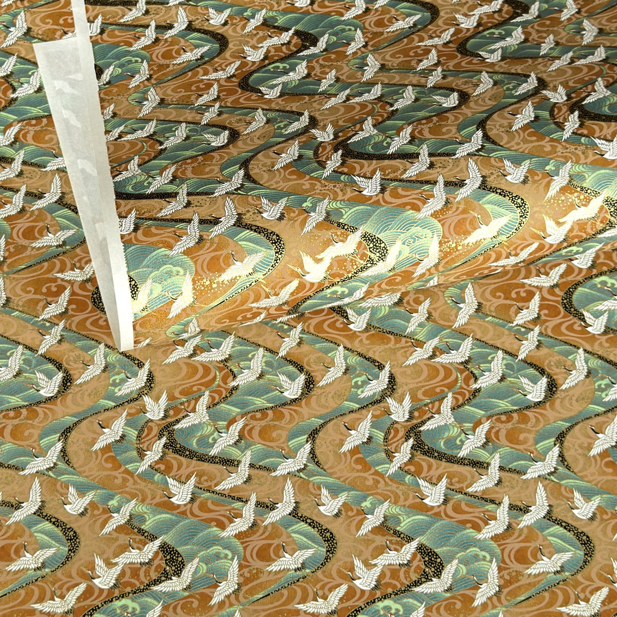 a sheet of Japanese chiyogami yuzen paper screen printed with a pattern of white flying cranes on a background of aqua and tan swirling lines