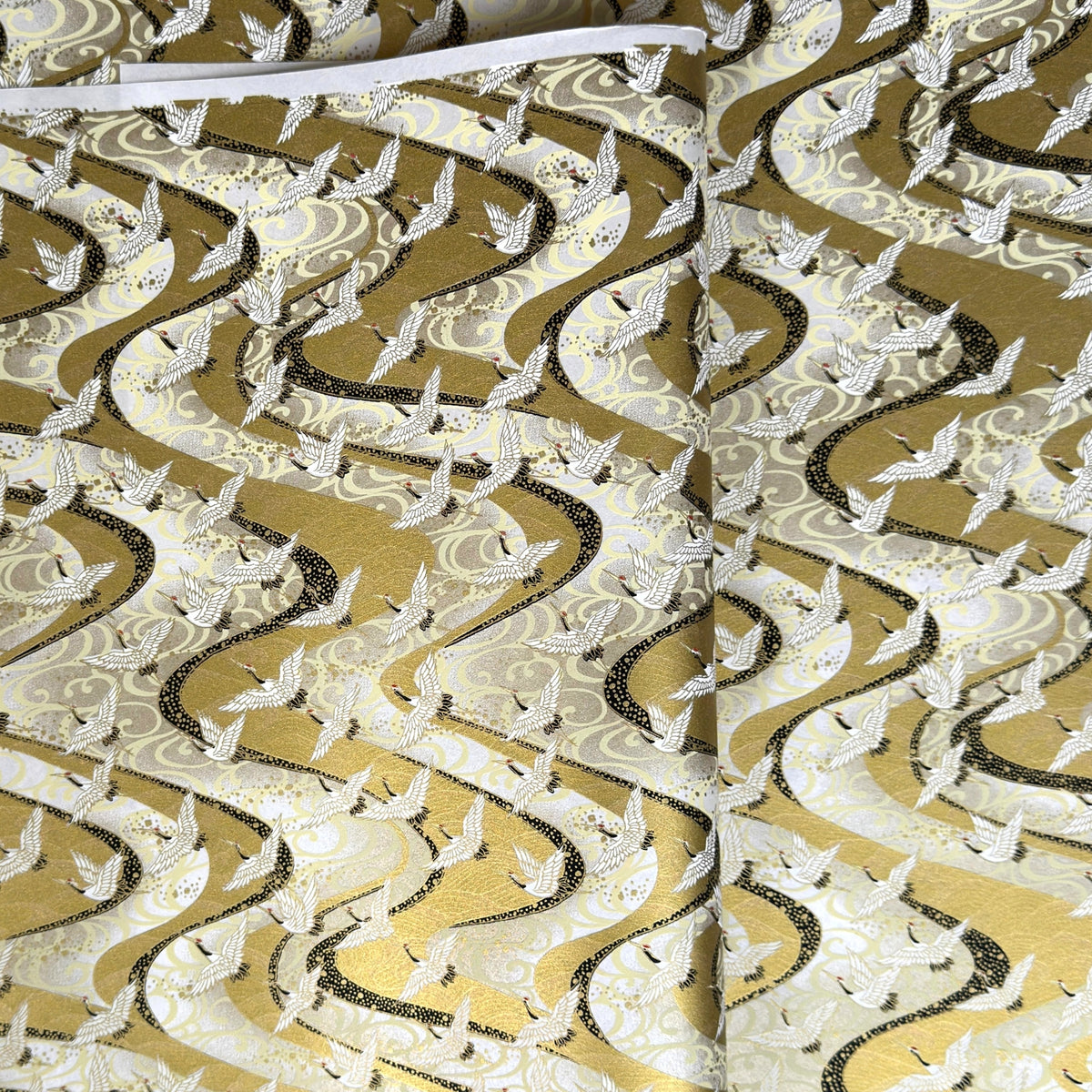 a sheet of Japanese chiyogami paper hand screen printed with a pattern of white cranes in flight.  The back drop is silver and lack swirling lines on gold. 