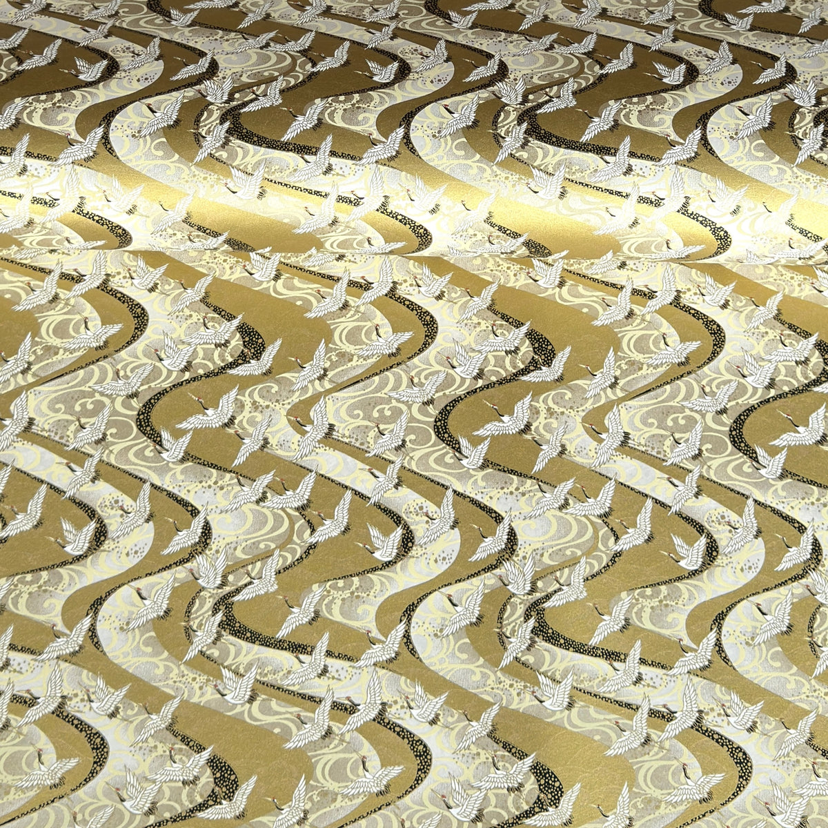 a sheet of Japanese chiyogami paper hand screen printed with a pattern of white cranes in flight.  The back drop is silver and lack swirling lines on gold. 