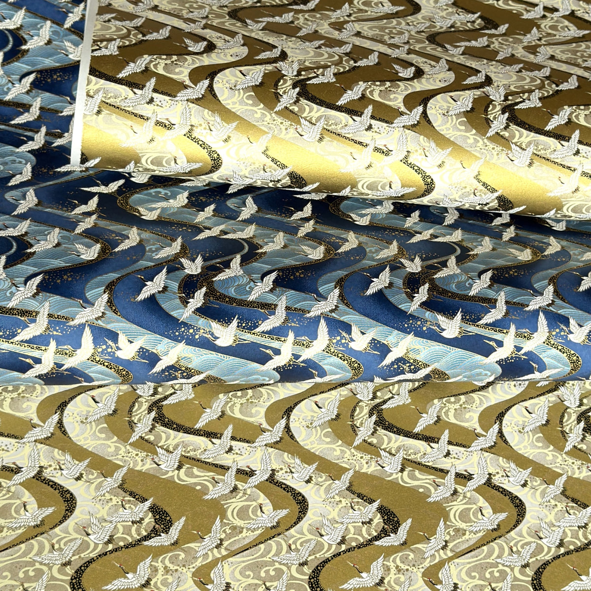 sheets of Japanese chiyogami paper with patterns of white cranes in flight on a backdrop of swirling lines.  The top of bottom papers pictured are in gold, the middle one is blue.