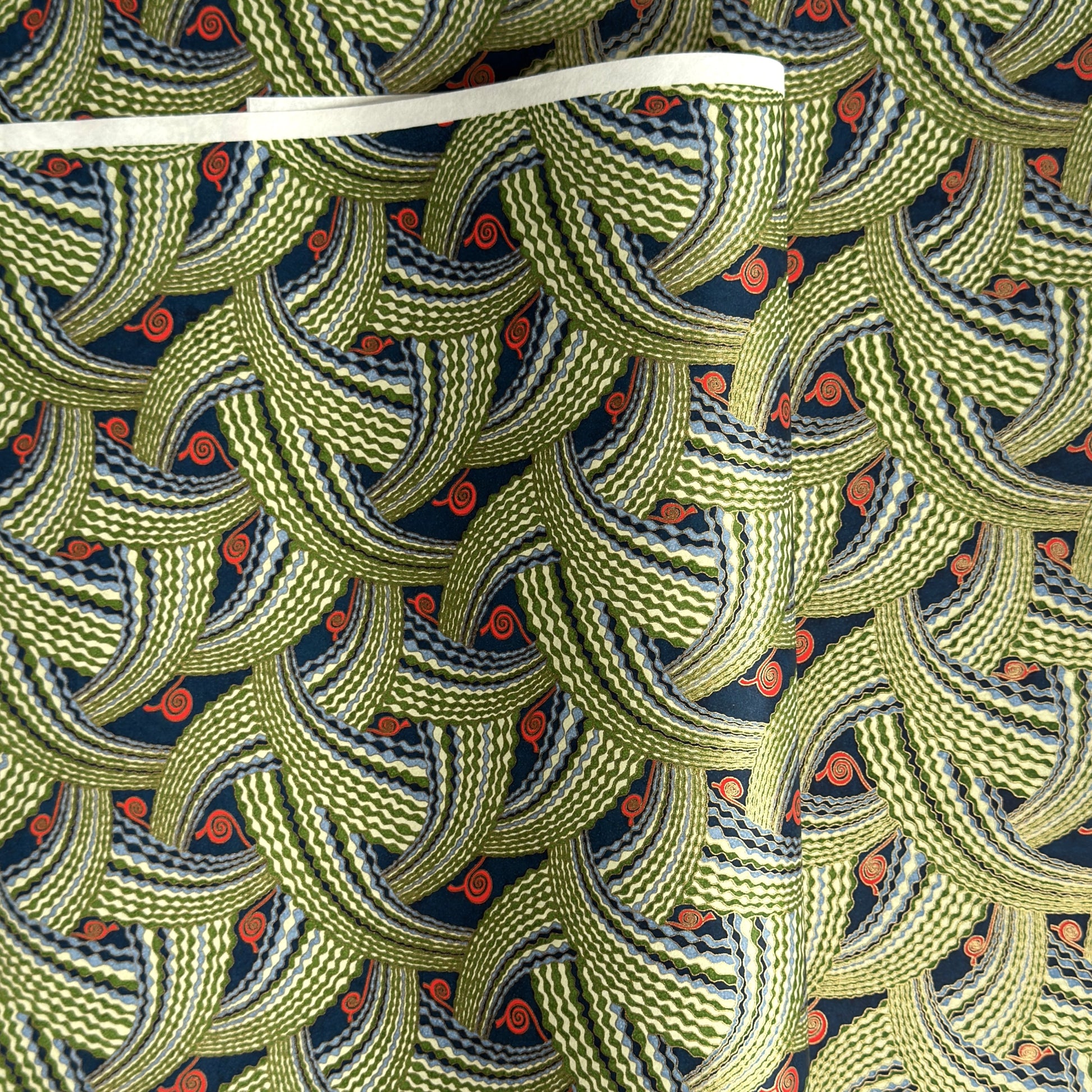 A close-up of a sheet of Japanese chiyogami paper with repeating curved wave-like motifs in olive green, navy blue, white, and small orange-red accents. 