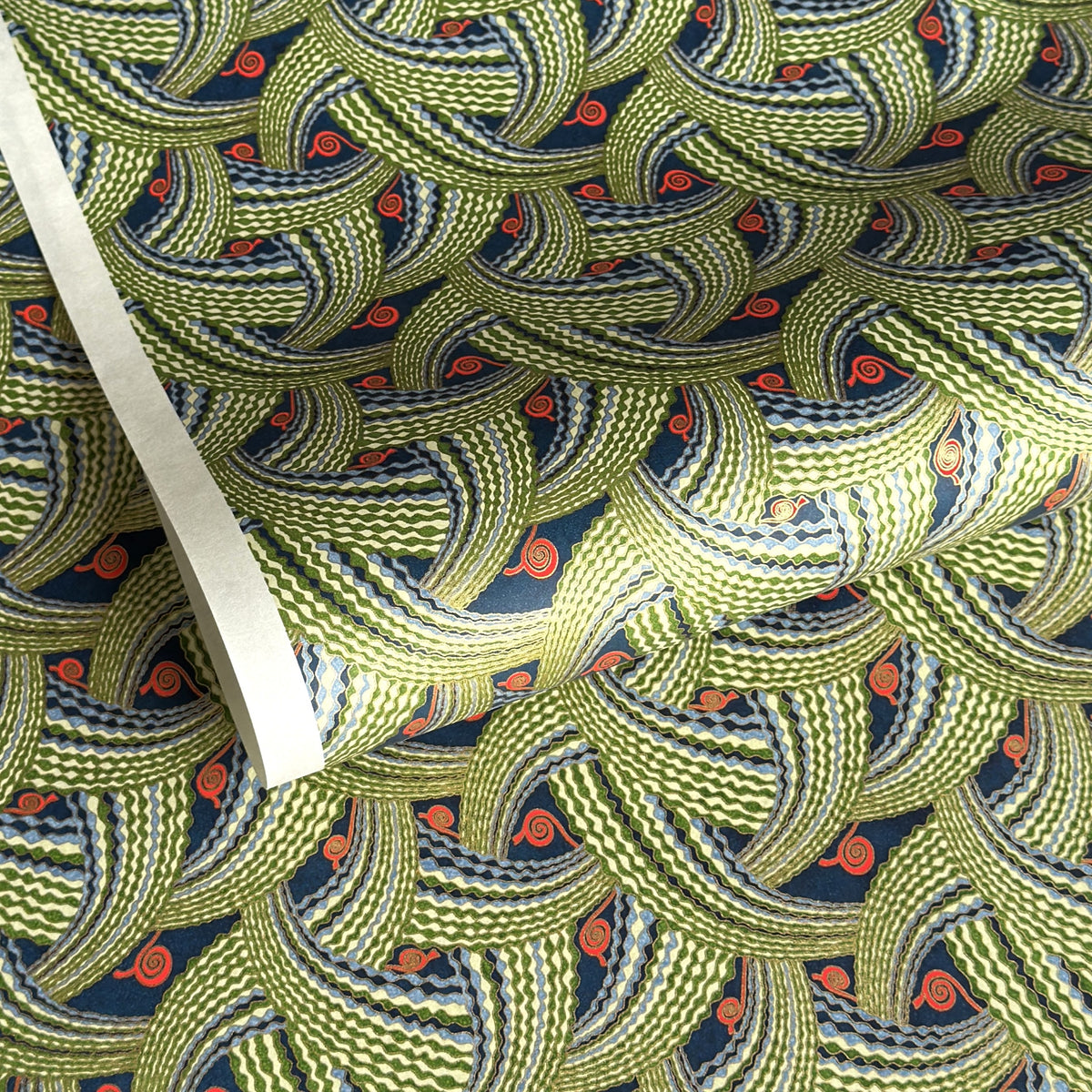 A close-up of a sheet of Japanese chiyogami paper with repeating curved wave-like motifs in olive green, navy blue, white, and small orange-red accents. 