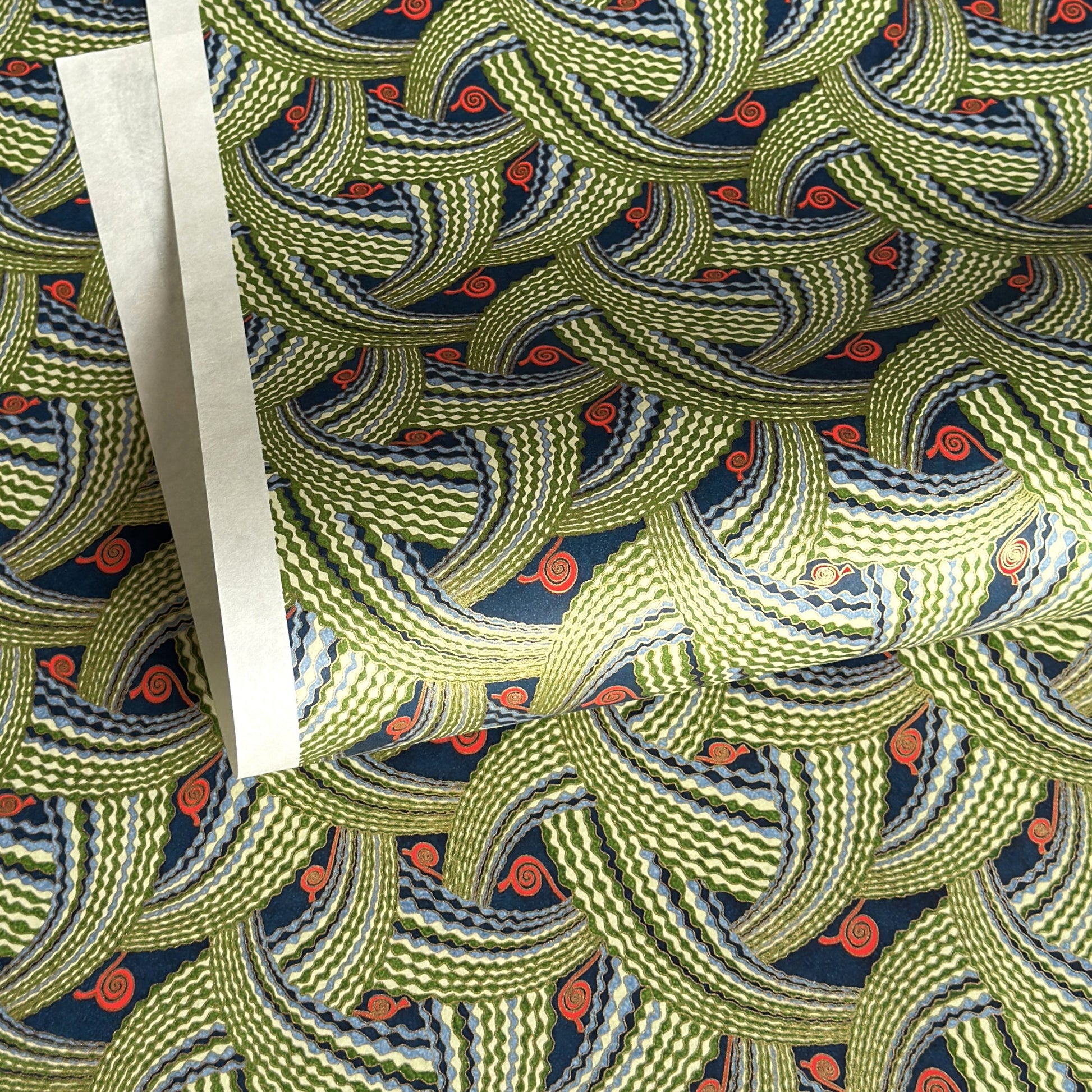 A close-up of a sheet of Japanese chiyogami paper with repeating curved wave-like motifs in olive green, navy blue, white, and small orange-red accents. 