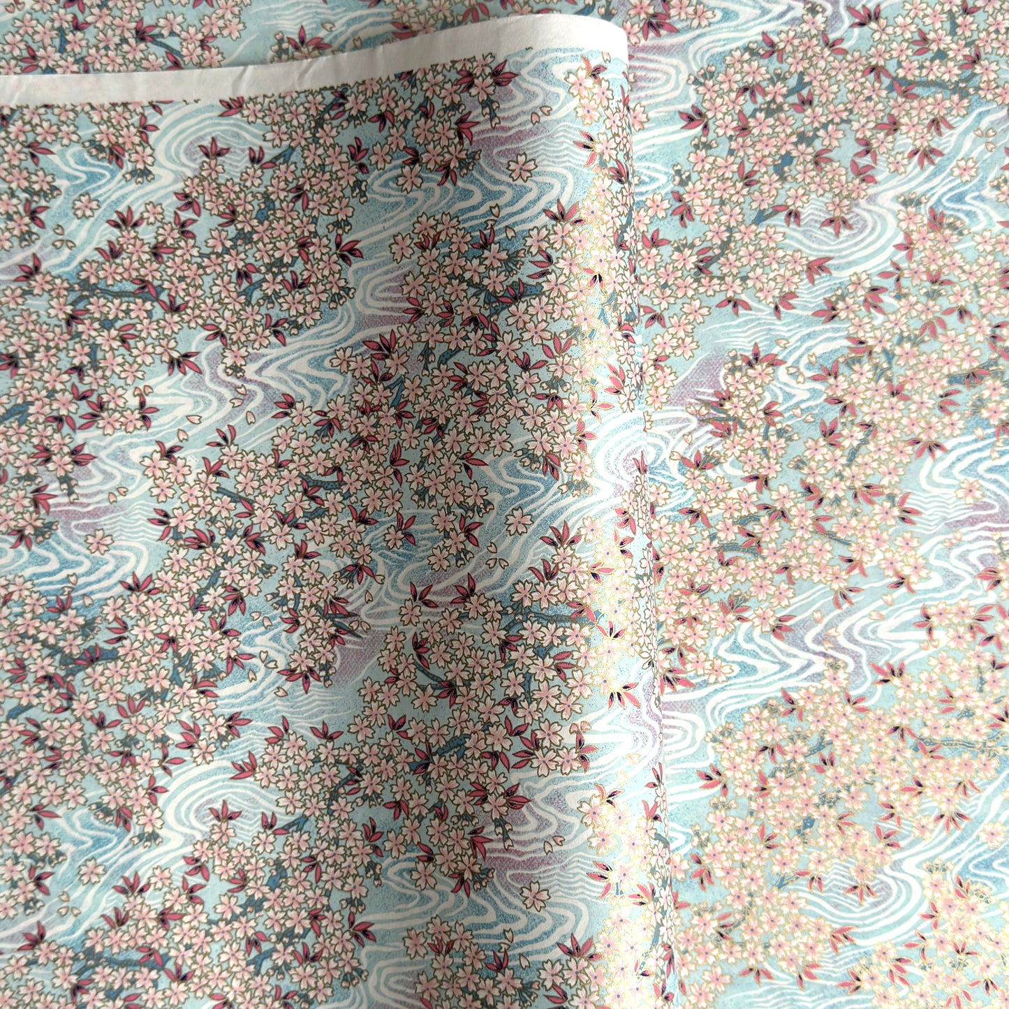 Japanese silkscreen chiyogami paper with an abundance of pale pink cherry blossom with a pale blue backdrop of meandering water, finished with metallic gold ink.