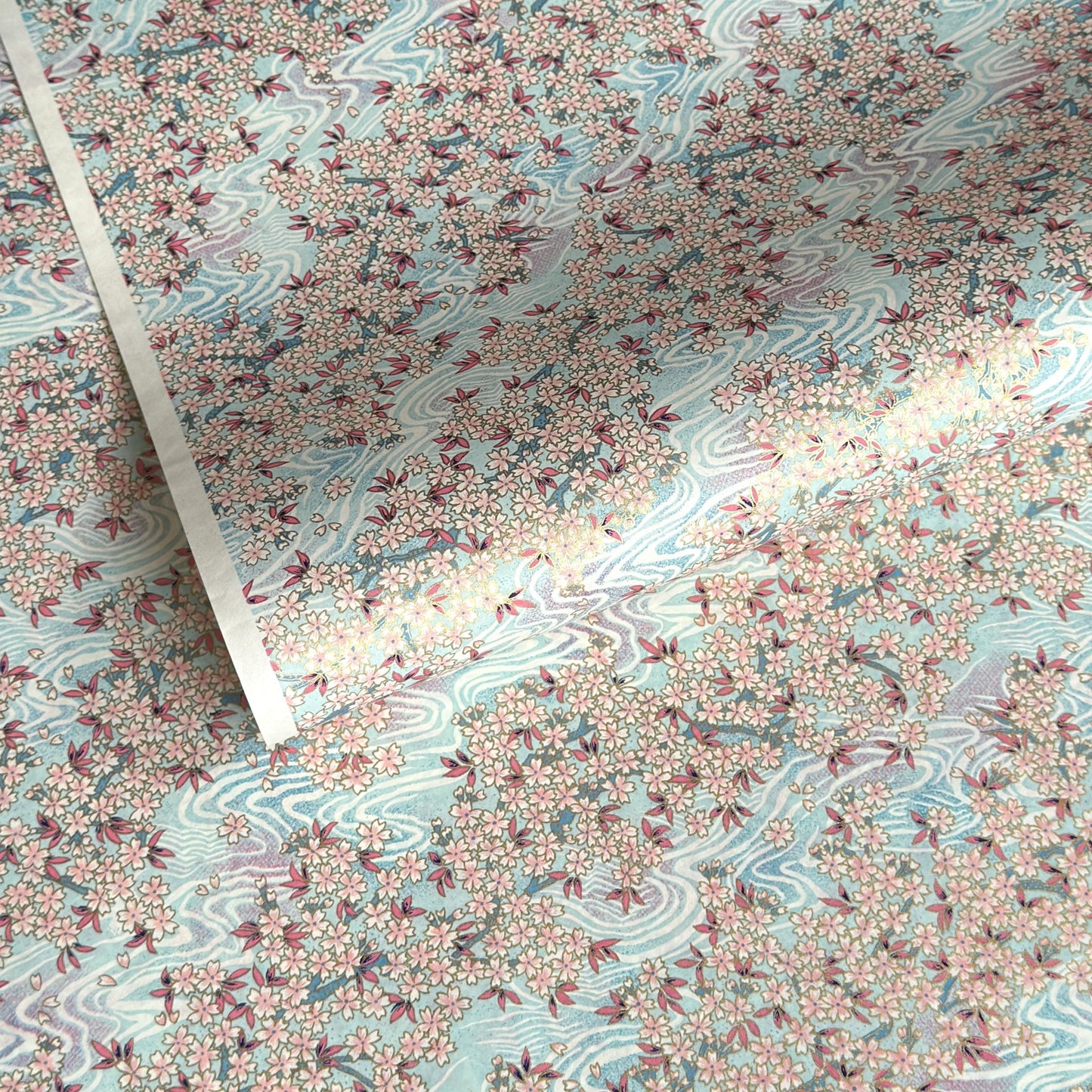 Japanese silkscreen chiyogami paper with an abundance of pale pink cherry blossom with a pale blue backdrop of meandering water, finished with metallic gold ink.