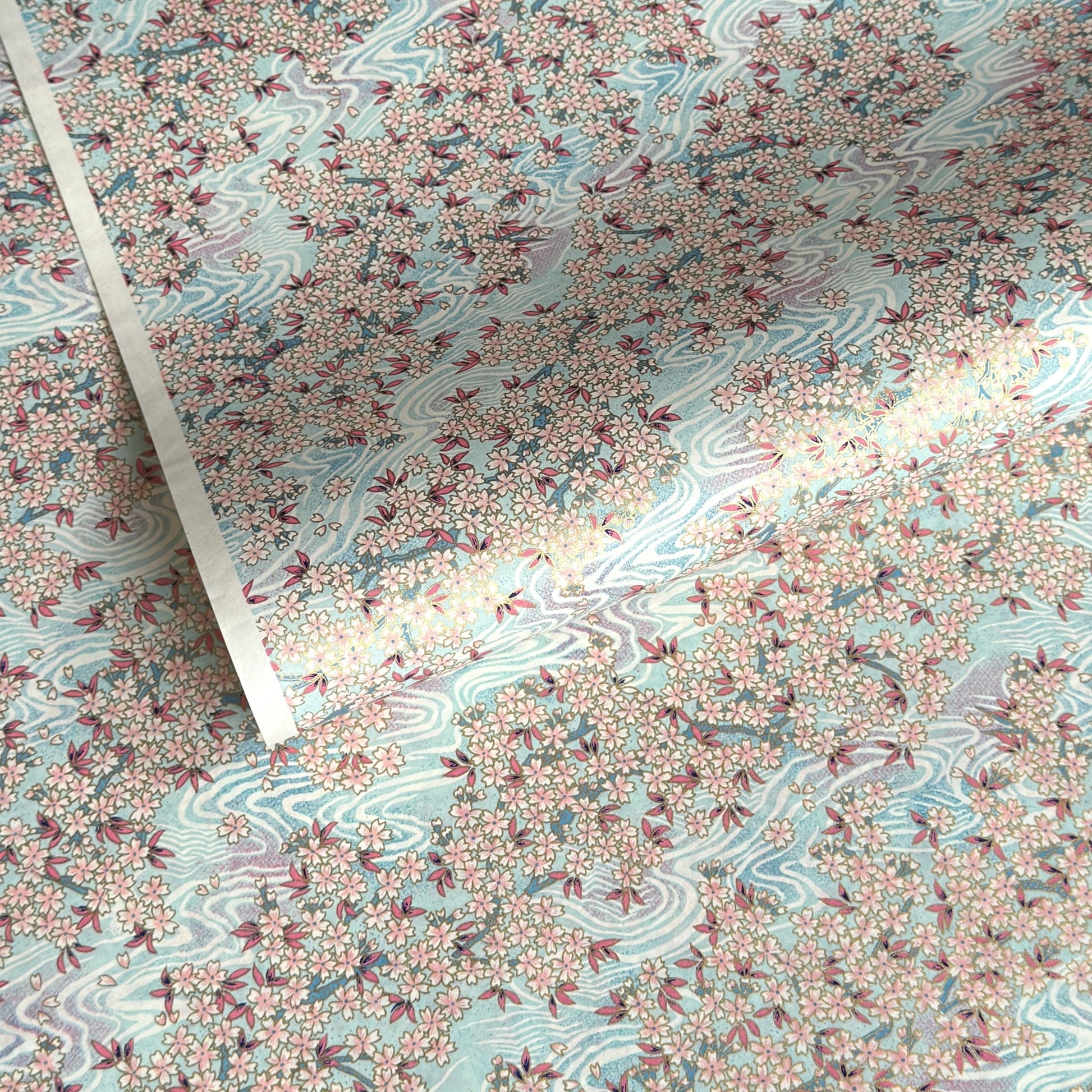 Japanese silkscreen chiyogami paper with an abundance of pale pink cherry blossom with a pale blue backdrop of meandering water, finished with metallic gold ink.