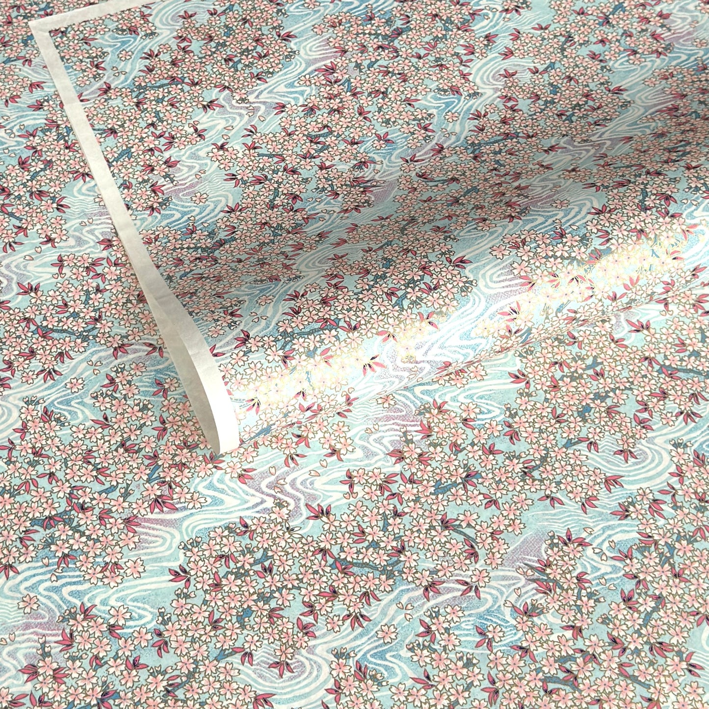 Japanese silkscreen chiyogami paper with an abundance of pale pink cherry blossom with a pale blue backdrop of meandering water, finished with metallic gold ink.