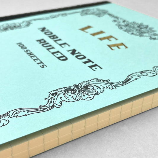 Softcover notebook with lined pages. The cover is plain aqua with a decorative black border and branding, Life Noble Ruled by Japanese brand Life