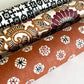 A Japanese stencil-dyed patterned paper with a repeat pattern of floral motif circles on a rich tan brown base. Rolled alongside other patterns.