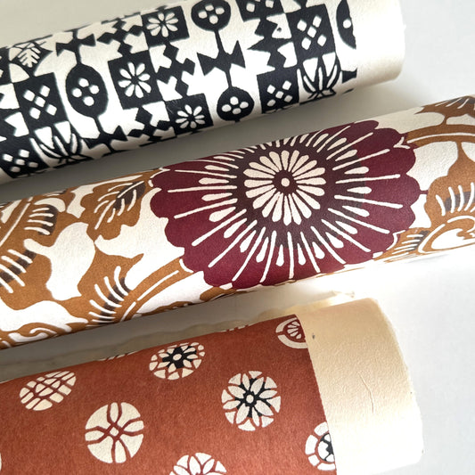 Japanese stencil-dyed paper with a large stylised floral pattern of a chrysanthemum in deep brown and tan. Pictured rolled with other paper