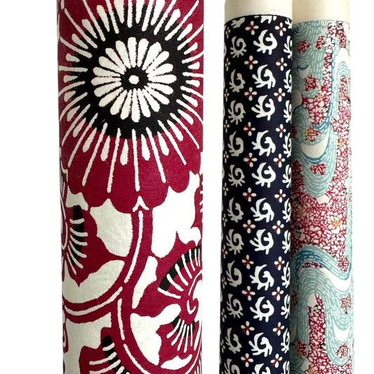 Japanese stencil-dyed paper with a repeat pattern of chrysanthemum flowers and swirling vines in deep berry red.  Pictured rolled with other patterned designs