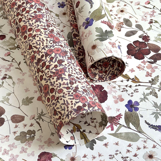 a sheet of wrapping paper with a pretty floral design of pressed flowers on a white background. Pictured rolled.  The reverse of the paper has a small scale rose design