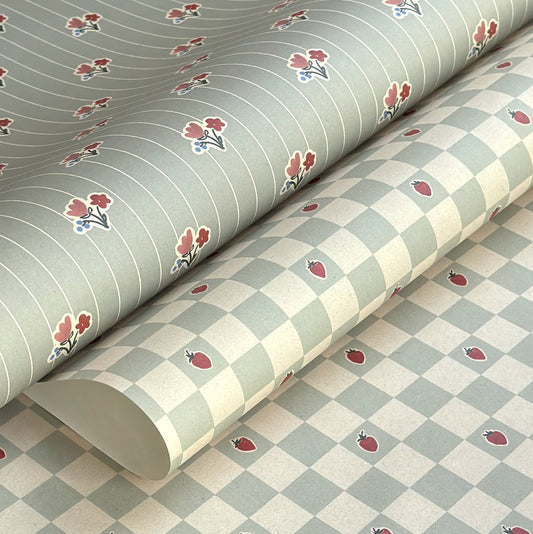 a sheet of wrapping paper with a soft green and cream checkered pattern with vintage style strawberries. Pictured alongside a stripe from the same collection