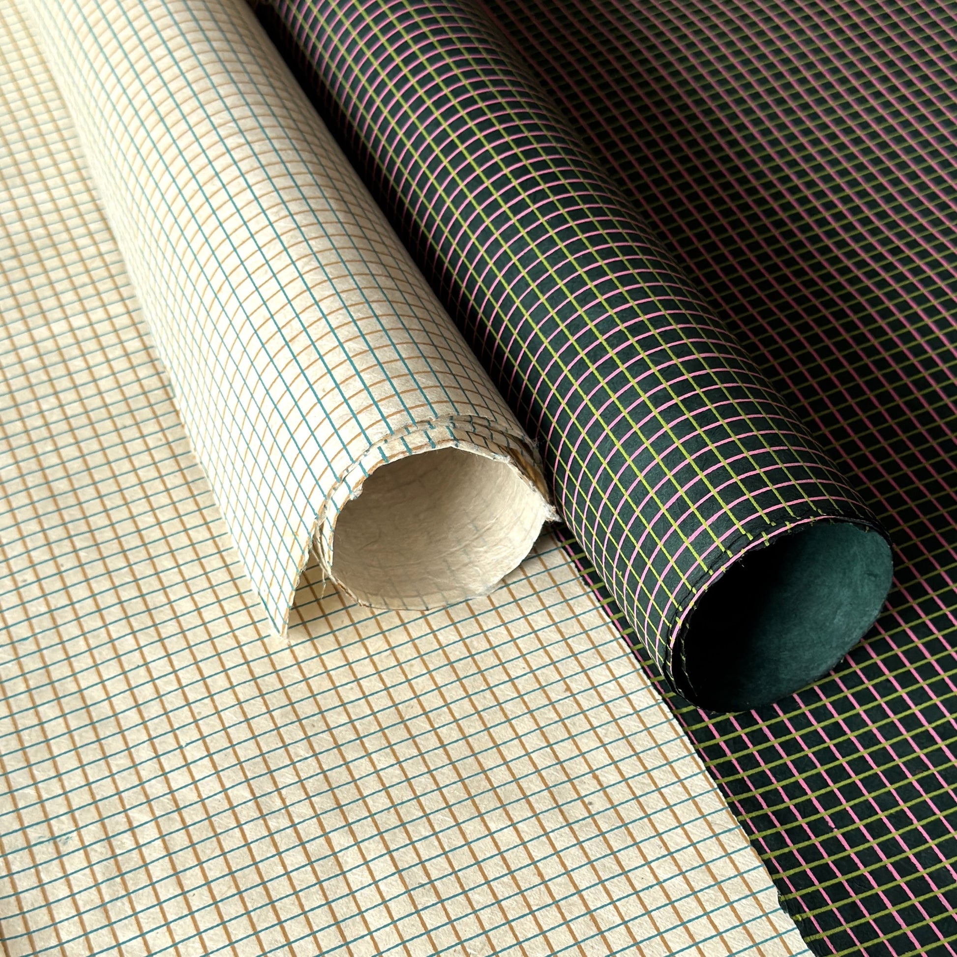 a sheet of Nepalese lokta paper with a lime green and pink grid pattern on dark green handmade paper. Pictured rolled alongside an ivory grid paper 