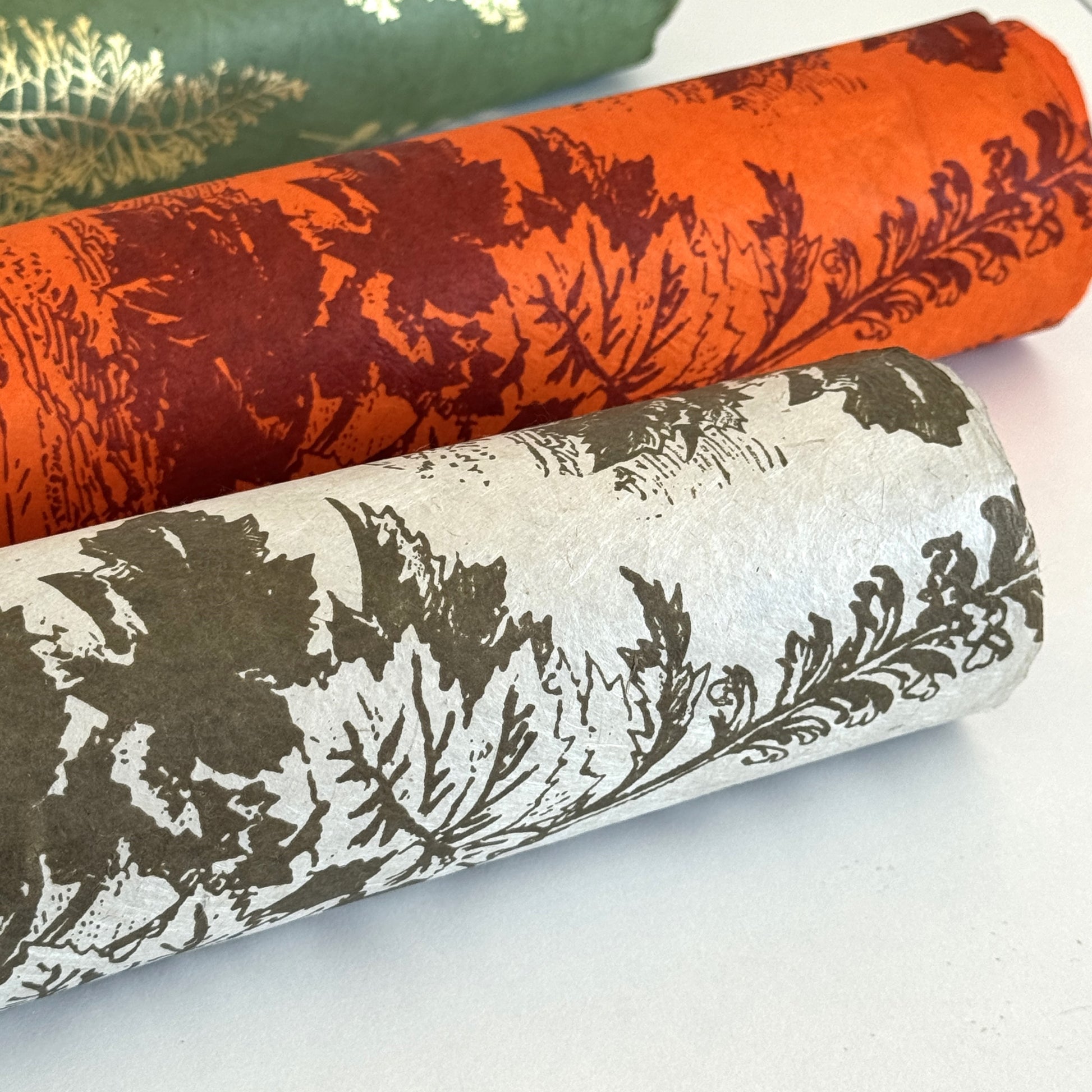 Nepalese handmade decorative lokta paper sheet with a Toile de Jouy style pattern of an agapanthus flower in burgundy screen printed on an orange lokta paper. Rolled with other designs