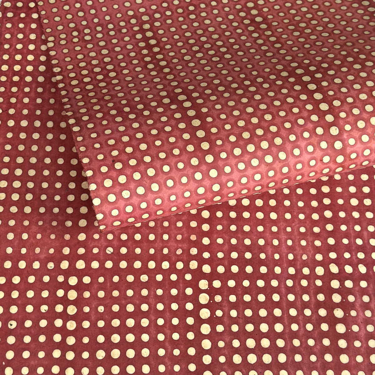 a sheet of Nepalese batik lokta paper in coral rose colour with a repeat white dot design. Pictured lightly rolled