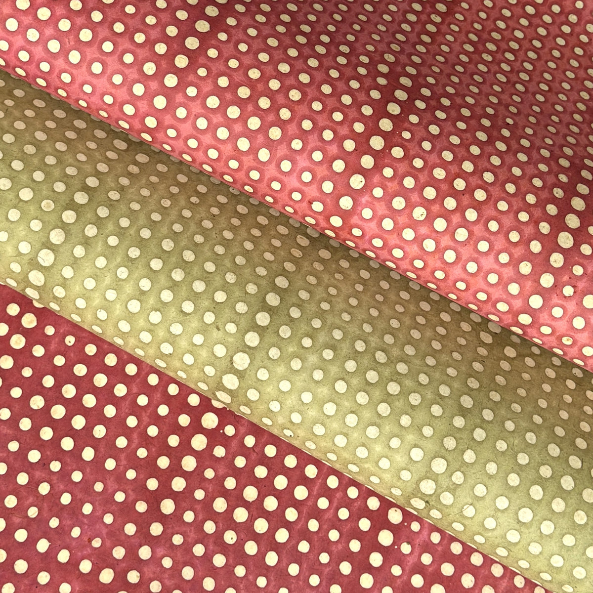 a sheet of Nepalese batik lokta paper in coral rose colour with a repeat white dot design. Pictured lightly folded alongside a sheet of the same design in sage green
