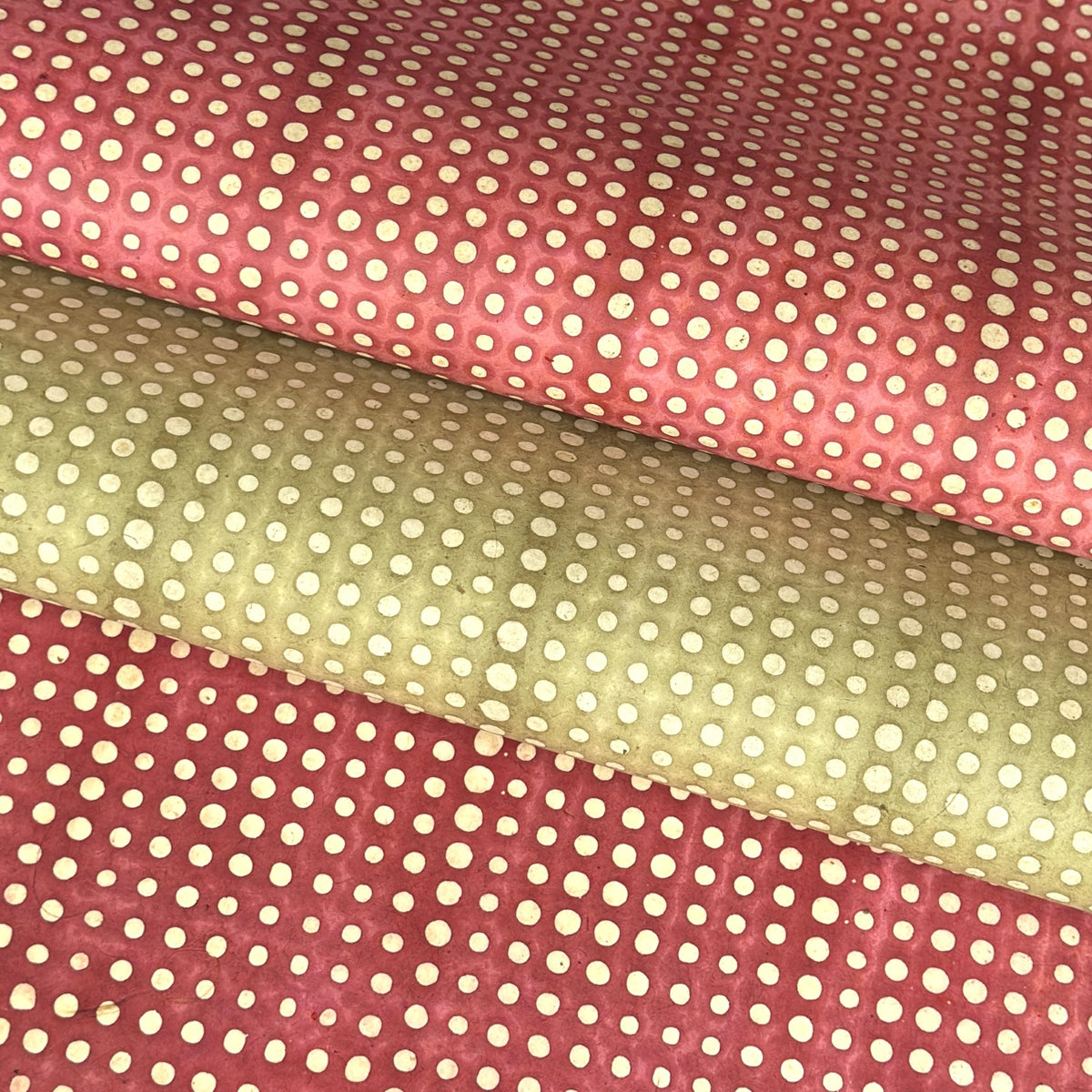 a sheet of Nepalese batik lokta paper in sage green with a repeat white dot design. Pictured lightly rolled with another paper in the red version of the same design