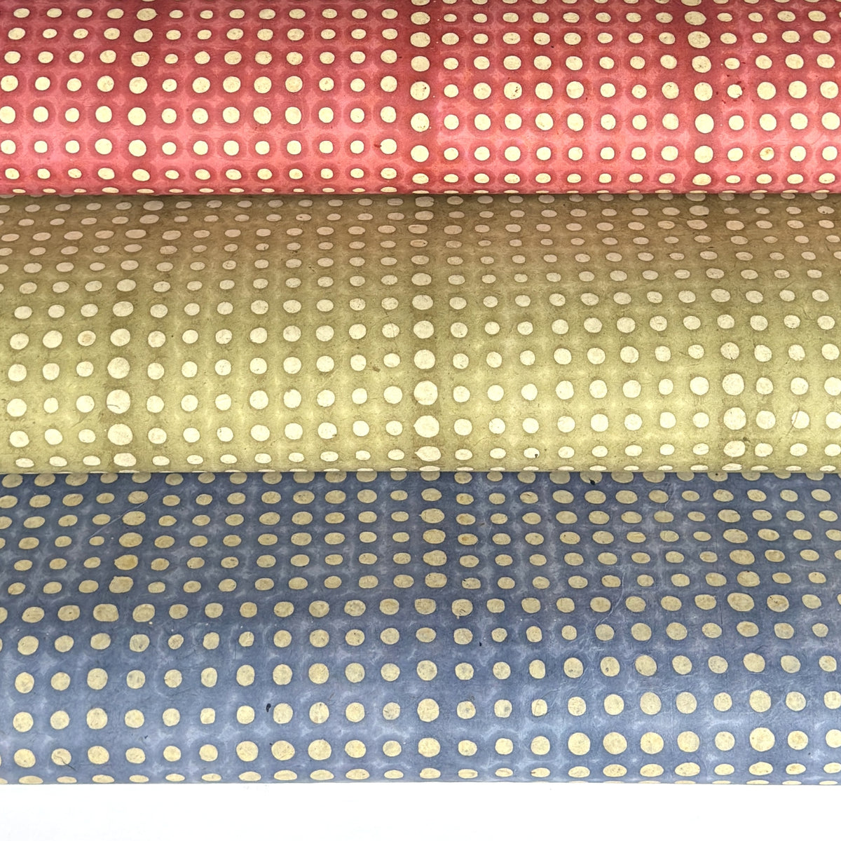 a sheet of Nepalese batik lokta paper in sky blue with a repeat white dot pattern.  Pictured rolled alongside the same designs in sage green and coral rose