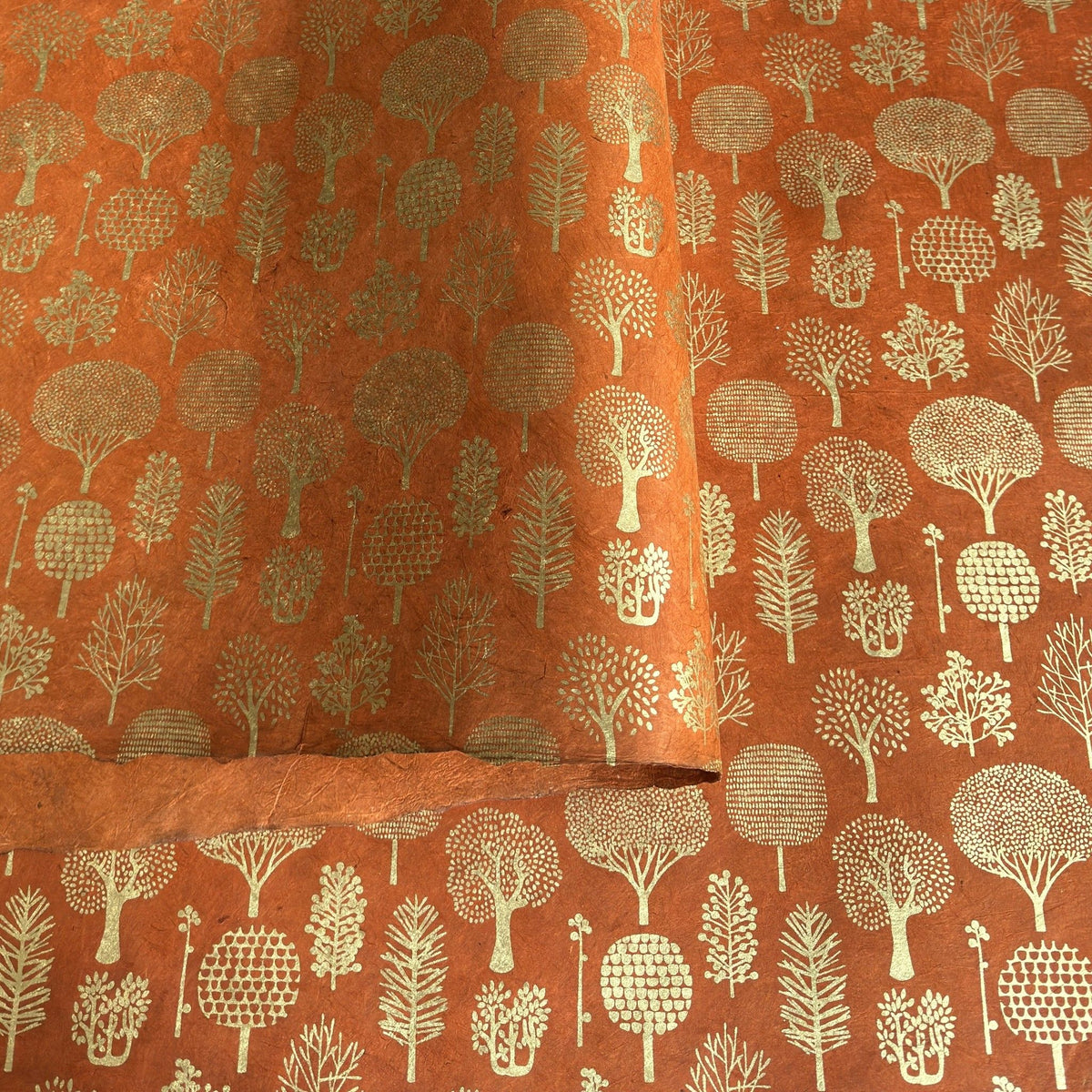 a sheet of screen printed Nepalese lokta paper in terracotta brown with gold trees.  The sheet is pictured lightly folded to show the gold of the trees and the deckled edge of the paper.