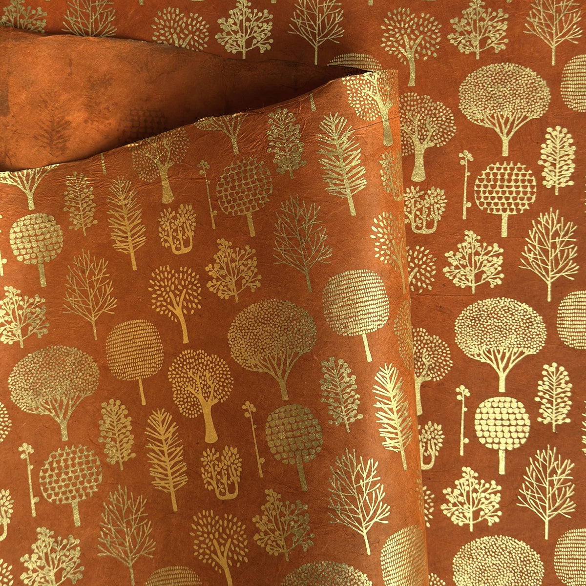 a sheet of screen printed Nepalese lokta paper in terracotta brown with gold trees.  The sheet is pictured lightly folded to show the gold of the trees and the deckled edge of the paper.