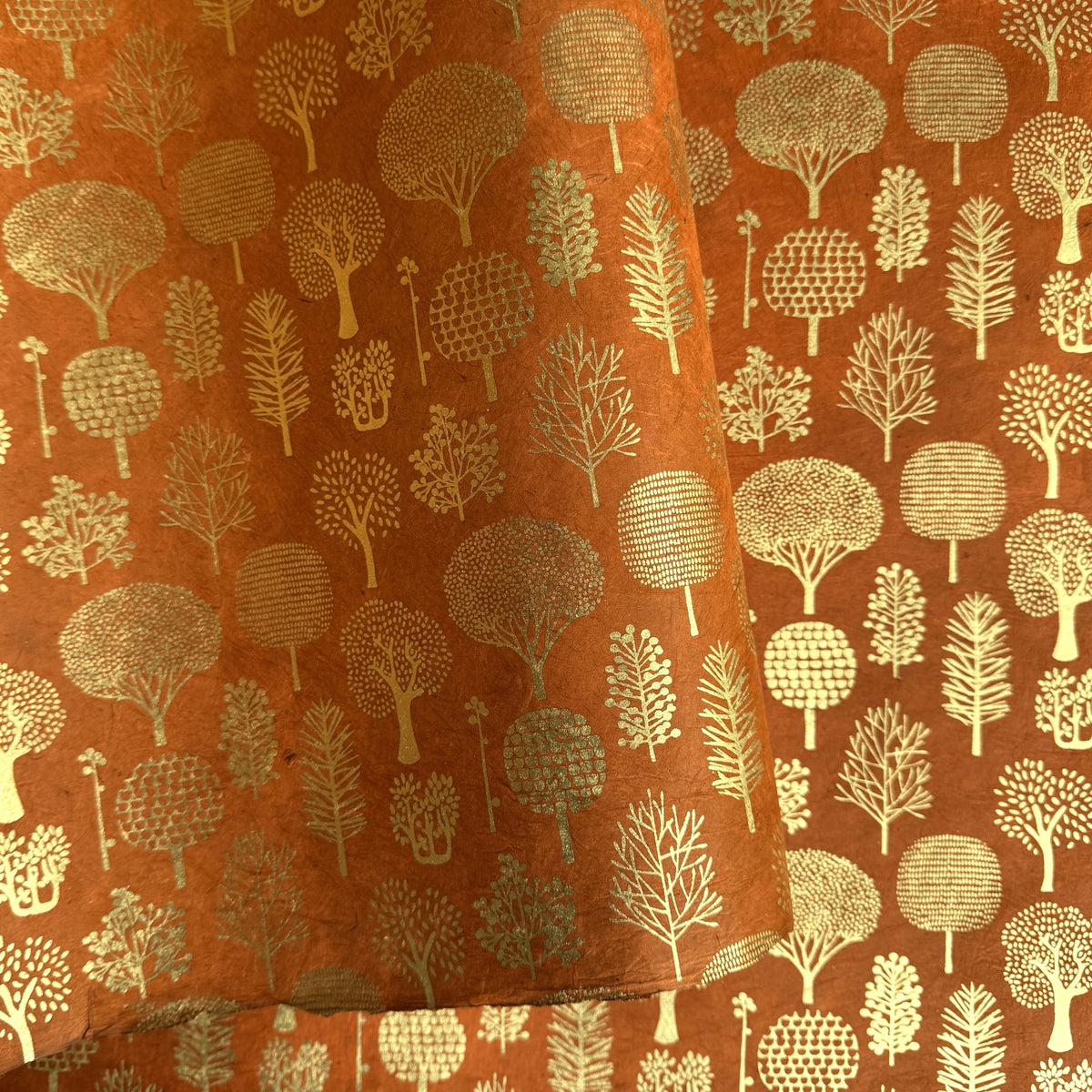 a sheet of screen printed Nepalese lokta paper in terracotta brown with gold trees.  The sheet is pictured lightly folded to show the gold of the trees and the deckled edge of the paper.