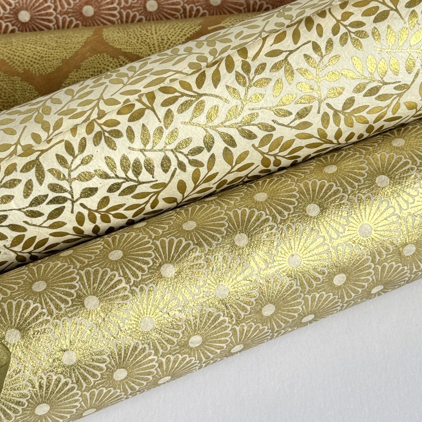 Nepalese handmade decorative lokta paper sheet with a gold metallic repeat floral pattern screen printed on an ivory lokta paper. Pictured rolled with other designs