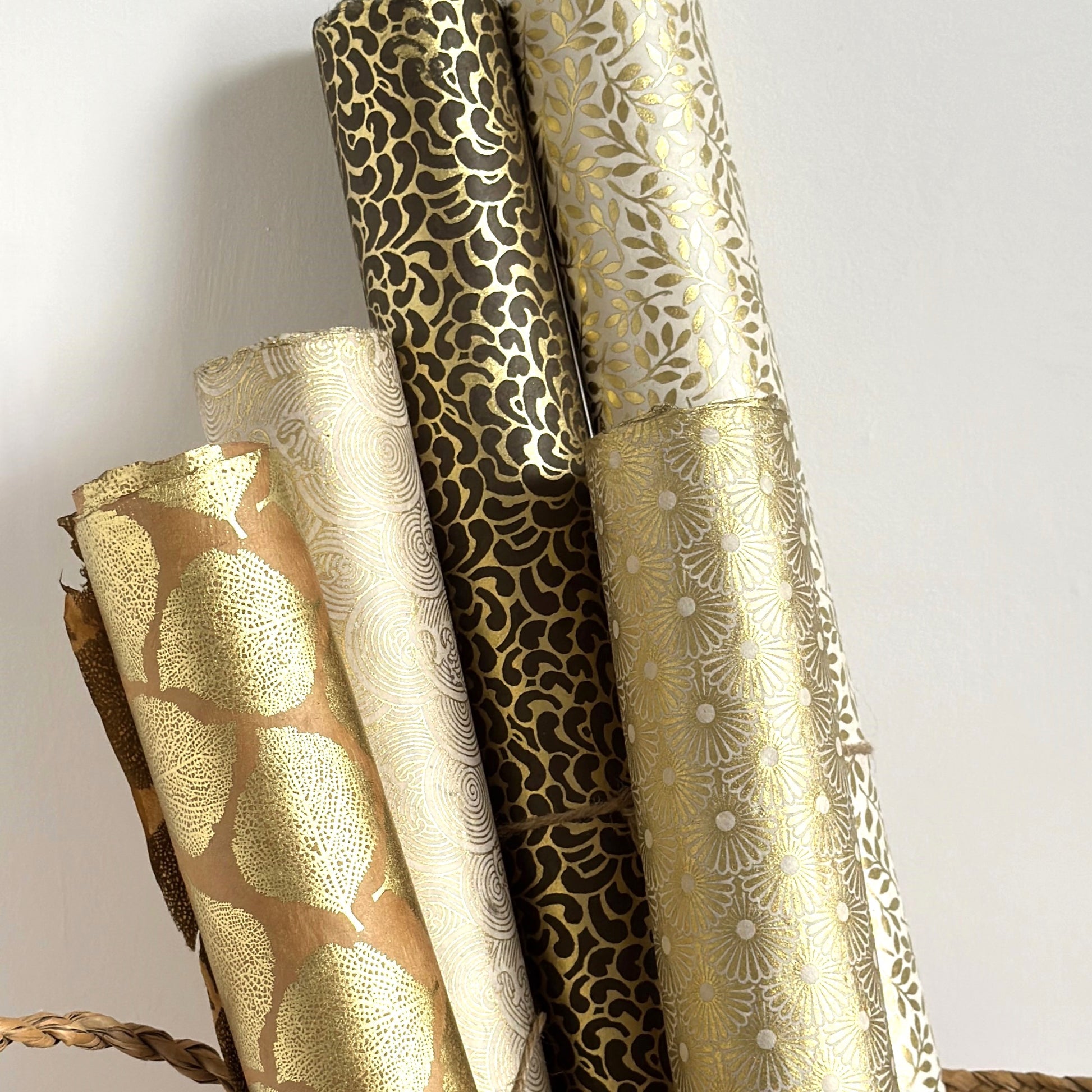 Nepalese handmade decorative lokta paper sheet with a gold metallic repeat floral pattern screen printed on an ivory lokta paper. Pictured rolled with other gold patterned designs.