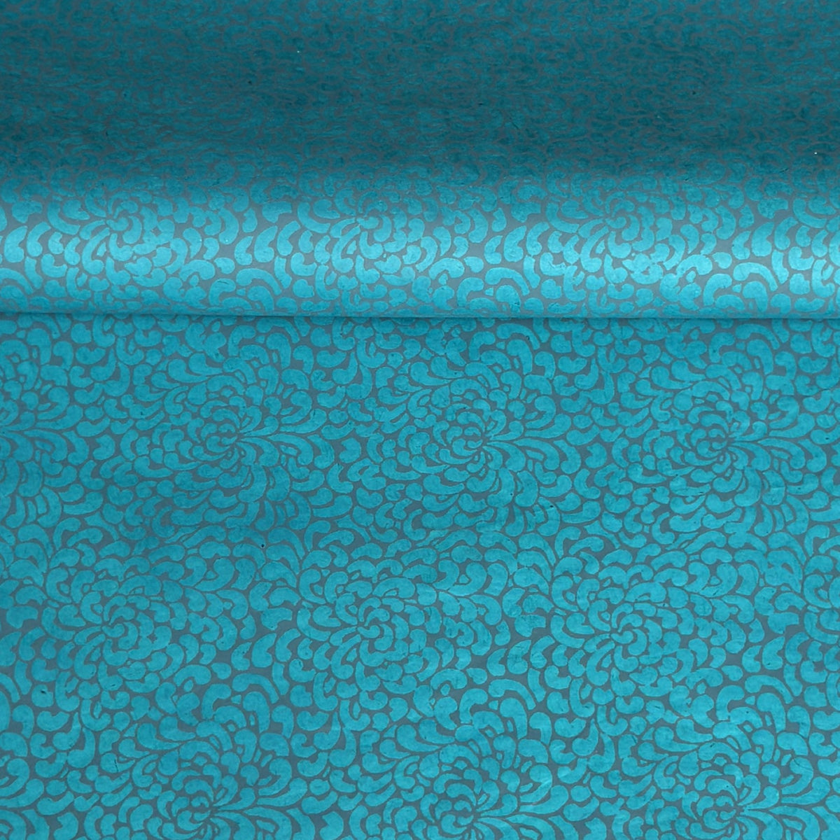 a sheet of Nepalese lokta paper screen printed with a repeat floral pattern in dark grey/blue on a bright teal lokta paper