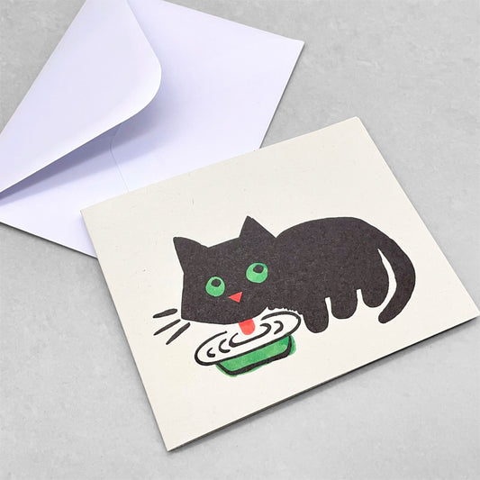 Small greetings card of a small black cat with big green eyes drinking milk from a bowl by Lisa Jones Studio