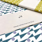 slimline exercise book with plain pages by Ola Studio, cover is a geometric repeat design in teal and lilac on a white background. Pictured with branded presentation band, close-up