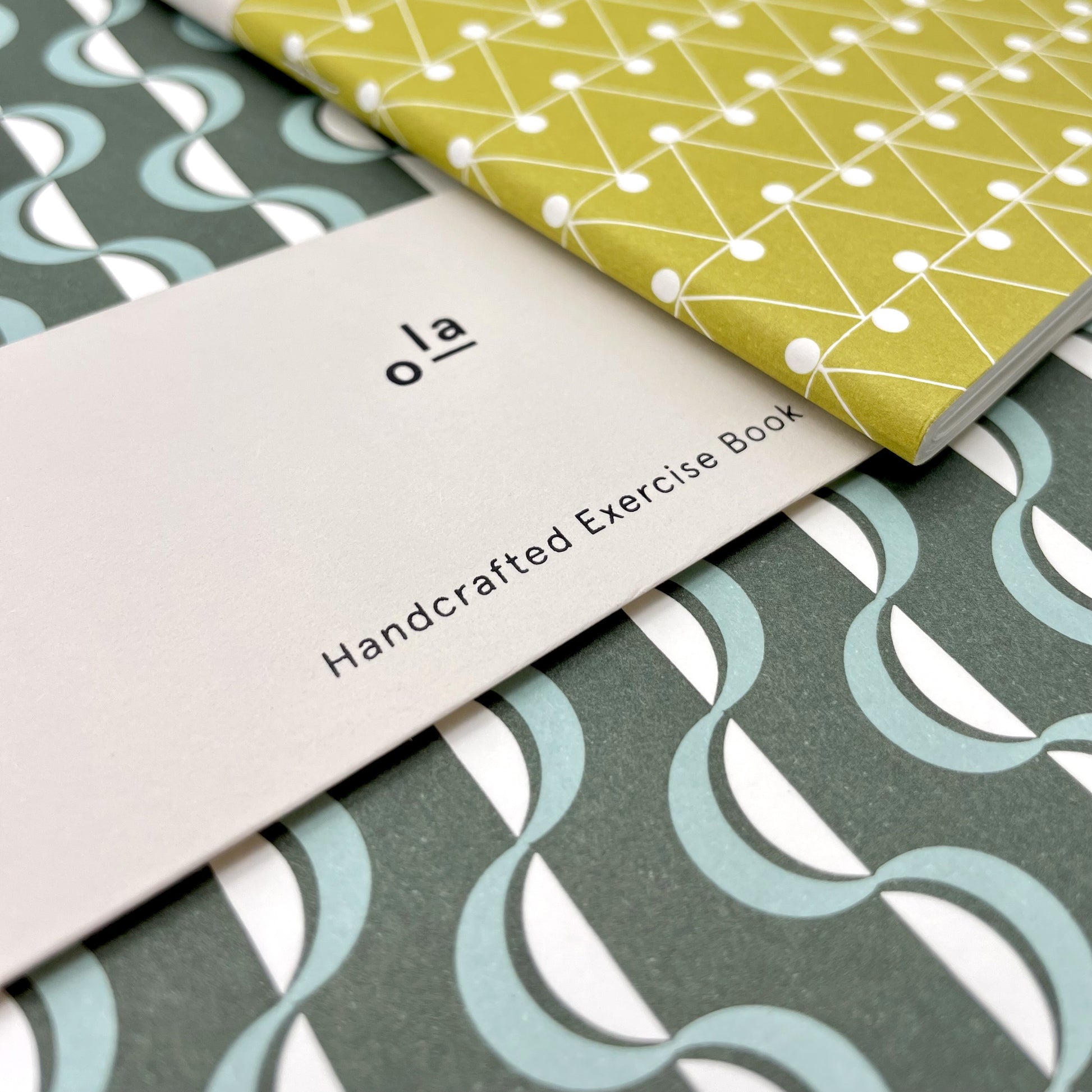 slimline softcover exercise book by Ola Studio, with plain pages. Front cover is a wavy stripe design in dark green and aqua on white. Pictured with branded presentation band, close-up
