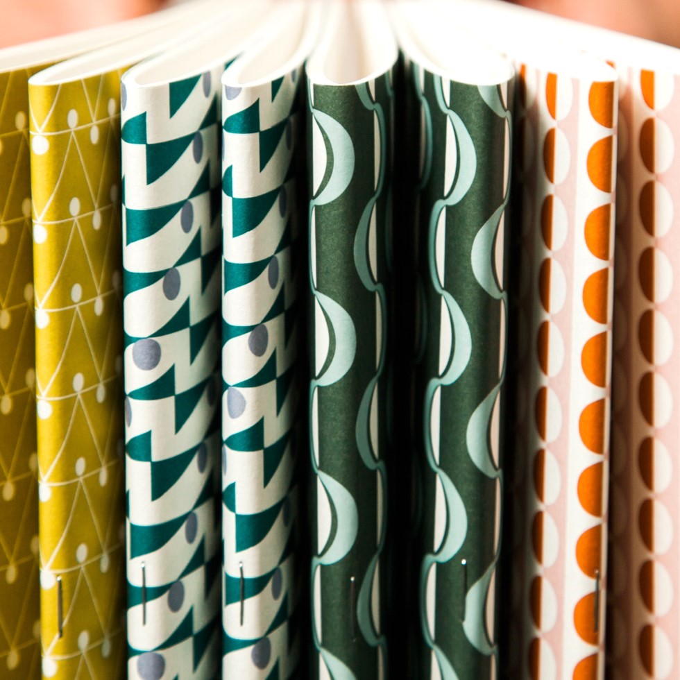 slimline softcover exercise book by Ola Studio, with plain pages. Front cover is a wavy stripe design in dark green and aqua on white. Pictured with other notebook designs from the collection