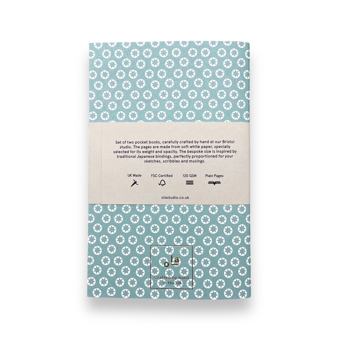 A set of two handcrafted pocket size notebooks by Ola Studio. One cover is a mustard and white triangle geometric design and the other is white stars on an aqua background. Reverse of the packaging